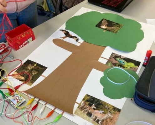 Plakat, auf das ein großer Baum aus buntem Papier sowie Fotos von Tieren aufgeklebt sind. Das Plakat ist über Kabel mit einem Makey Makey verbunden.