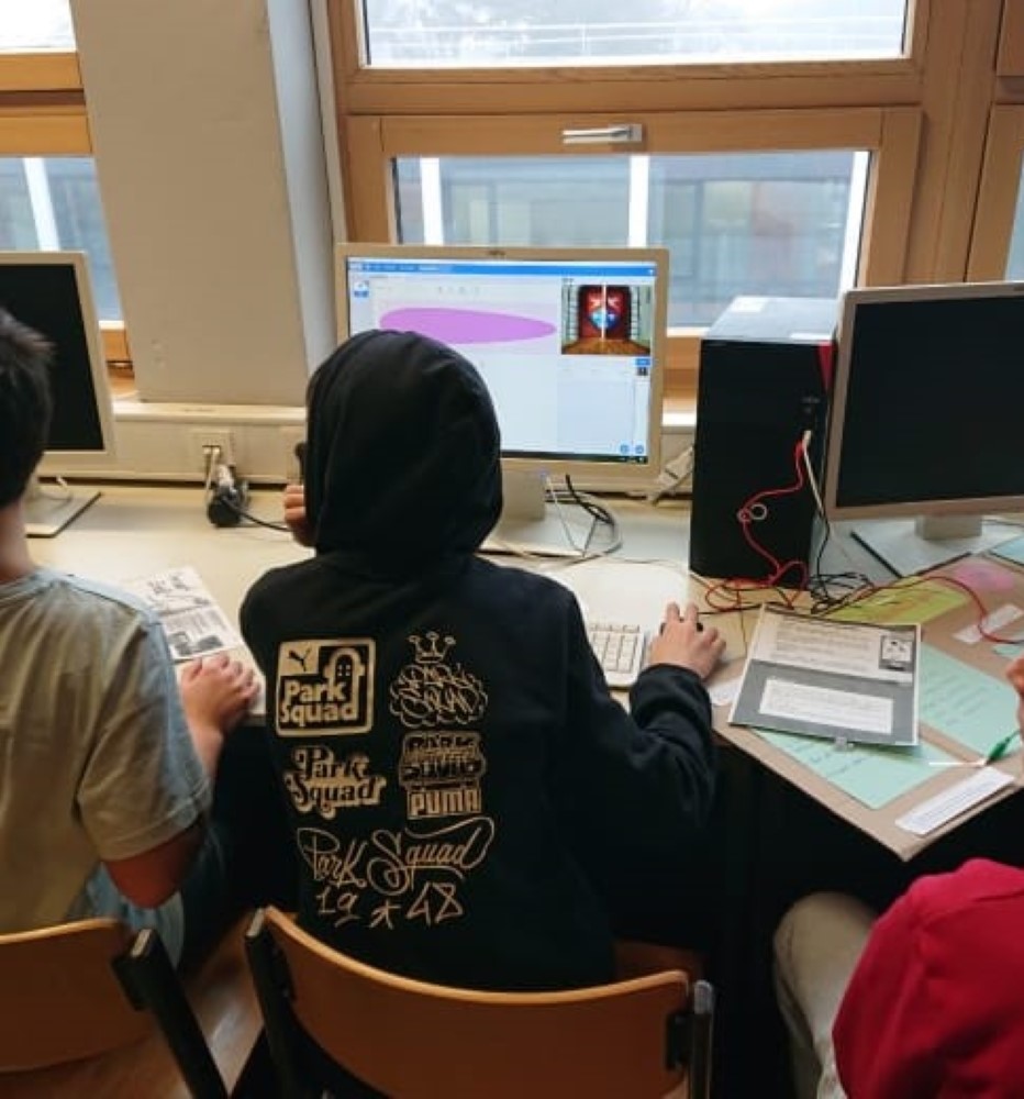 Drei Siebtklässler schauen auf einen Computer. Auf dem Tisch liegt ein Plakat, an dem sie gerade arbeiten.