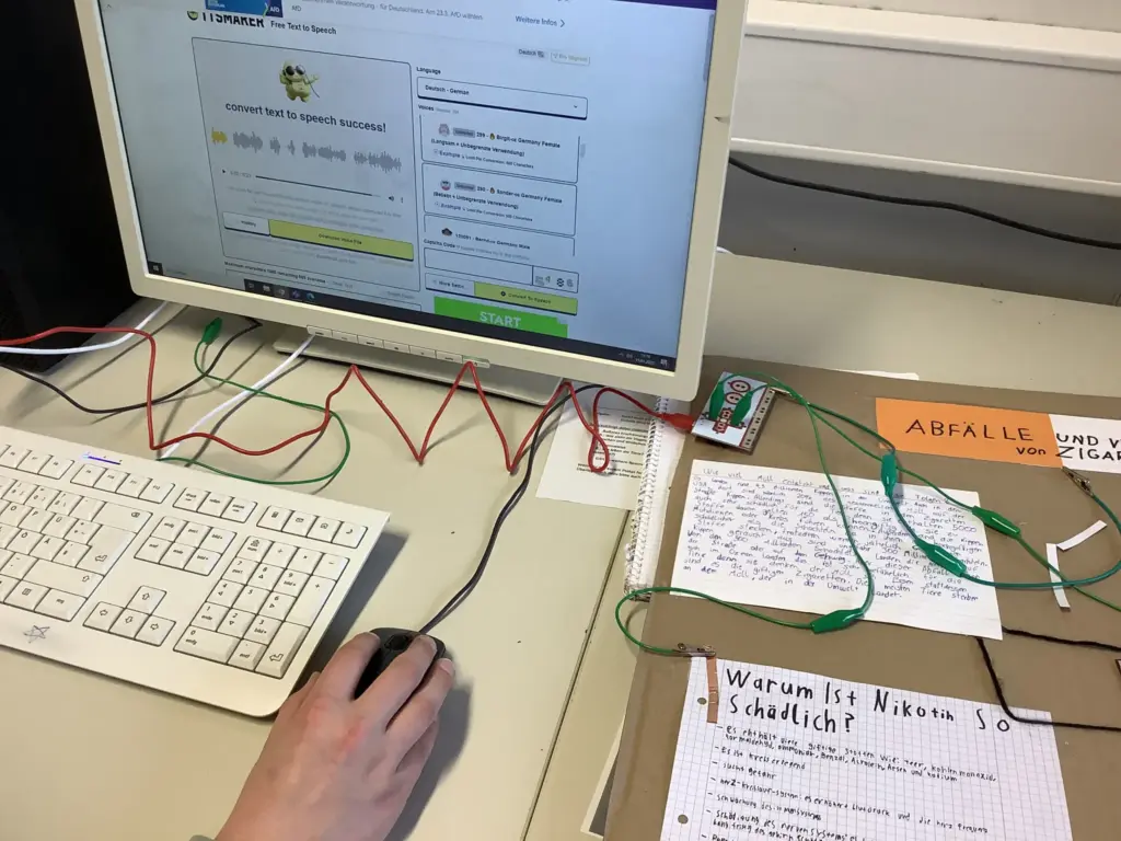 Es ist ein Computerbildschirm zu sehen, auf dem ein K I Modell Text in Sprache umwandelt. Daneben liegt ein Plakat zum Thema Rauchen und Nachhaltigkeit. Der Computer und das Plakat sind über Kabel verbunden.