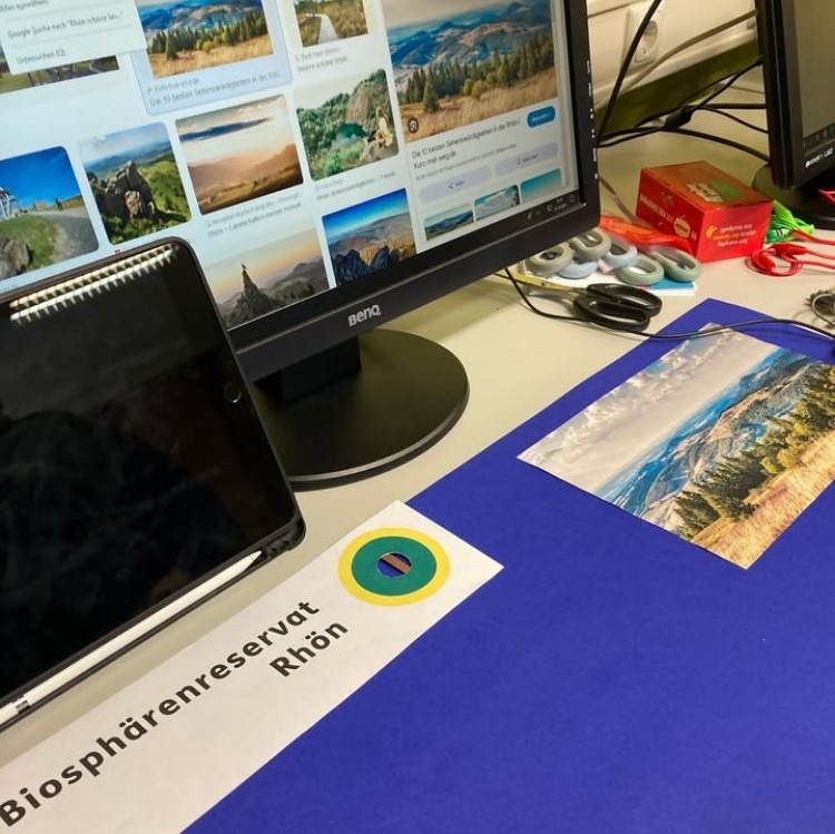Auf einem Tisch liegt ein Plakat mit der Überschrift Biosphärenreservat Rhön. Dahinter steht ein Computerbildschirm, auf dem Naturfotos zu sehen sind.