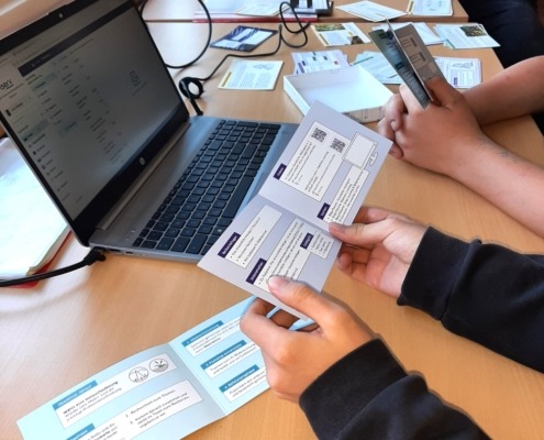 Auf drei aneinandergereihten Tischen liegen Karten und Spiele sowie zwei Laptops. Jugendliche sehen sich die Materialien an.