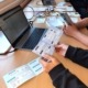 Auf drei aneinandergereihten Tischen liegen Karten und Spiele sowie zwei Laptops. Jugendliche sehen sich die Materialien an.