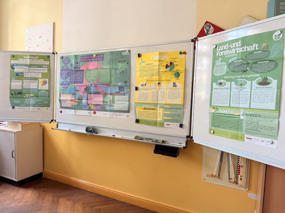An einem Whiteboard in einem Klassenraum hängen vier Plakate mit folgenden Titeln: Künstliche Intelligenz trifft natürlichen Klimaschutz. Land- und Forstwirtschaft. Deutsche und internationale Klimaziele. Und Energie. Sie sind aus den Programmen K I Box Klima und Klima Pakete