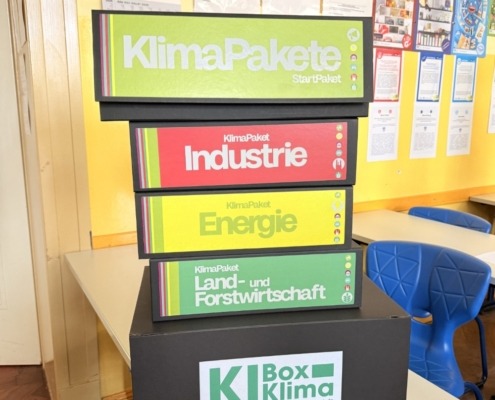 Fünf Materialboxen sind auf einem Tisch in einem Klassenraum aufeinander gestapelt. Sie haben folgende Aufschriften: Klima Pakete Startpaket. Klimapaket Industrie. Klimapaket Energie. Klimapaket Land- und Forstwirtschaft. Und K I Box Klima.