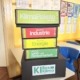 Fünf Materialboxen sind auf einem Tisch in einem Klassenraum aufeinander gestapelt. Sie haben folgende Aufschriften: Klima Pakete Startpaket. Klimapaket Industrie. Klimapaket Energie. Klimapaket Land- und Forstwirtschaft. Und K I Box Klima.