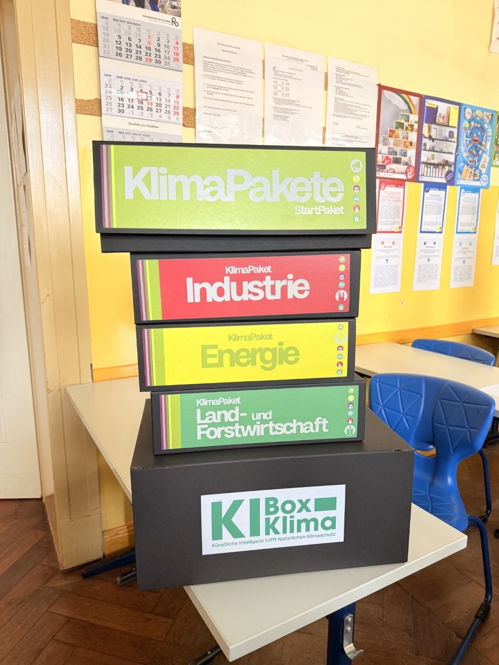 Fünf Materialboxen sind auf einem Tisch in einem Klassenraum aufeinander gestapelt. Sie haben folgende Aufschriften: Klima Pakete Startpaket. Klimapaket Industrie. Klimapaket Energie. Klimapaket Land- und Forstwirtschaft. Und K I Box Klima.