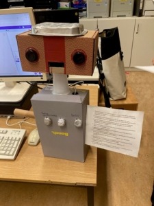Ein aus Kartons und Plastik gebauter Roboter steht auf einem Tisch vor einem Computer. An einem seiner Arme ist ein Blatt Papier mit gedrucktem Text angeklebt. Der Text ist auf dem Bild nicht zu erkennen.